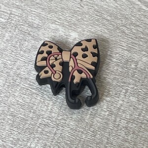 May include: A black shoe charm shaped like a bow with a leopard print pattern. A pink stethoscope is centered on the bow. The charm is designed to look like a bow with a stethoscope.