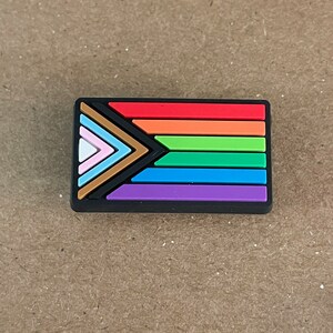 May include: A black and rainbow colored pride flag with a black triangle in the corner. The flag has seven stripes: brown, light blue, white, pink, orange, yellow, and green.