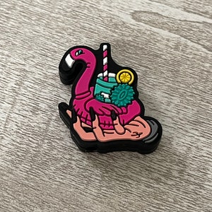 May include: A pink flamingo with a green drink and a yellow lemon slice on top. The flamingo is being held by two hands with pink skin and black fingernails.