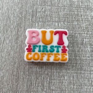 May include: A white sticker with the text "BUT FIRST COFFEE" in colorful letters. The letters are in a retro style with a coffee cup graphic on either side of the word "FIRST".