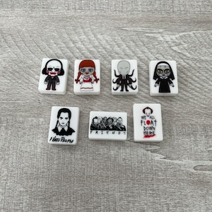 May include: Set of six white ceramic tiles with horror movie character illustrations. The tiles feature characters from the movies: Saw, Chucky, Slender Man, The Nun, Addams Family, and IT. The tiles are approximately 2 inches by 1 inch.
