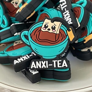 May include: A collection of teal, brown, and black "ANXI-TEA" charms. Each charm features a teal teacup with brown tea and a tea bag with a sad face. The charms are on a white surface.