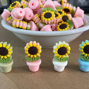 May include: Four miniature potted sunflowers with different colored pots. The pots are green, pink, white, and blue. The sunflowers have yellow petals and brown centers.