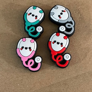 May include: Four colorful stethoscope charms with a cartoon face and red cheeks. The charms are made of silicone and are in the colors green, gray, pink, and red.
