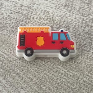 May include: A red toy fire truck with a yellow ladder, a white shield, and blue windows. The truck has black wheels and a white base.