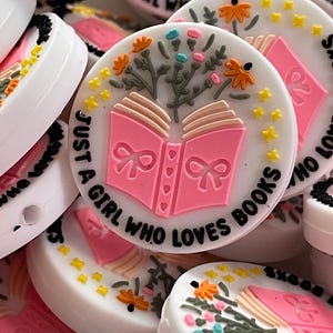 May include: A close-up of several white round silicone charms with pink and black text that reads "Just a girl who loves books". The charms feature a pink open book with a floral design and yellow stars.