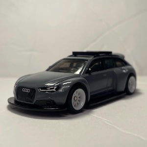 May include: A grey die-cast model car with white wheels. The car has a black roof rack and a black front spoiler. The front of the car features the Audi logo. The car is a miniature replica of a sports wagon.