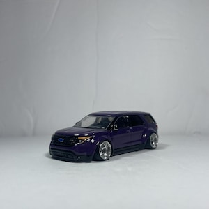 Peut inclure: Une voiture jouet SUV violette avec des jantes argentées et un pare-chocs avant noir. La voiture jouet a une suspension abaissée et est posée sur un fond blanc. Les phares de la voiture sont jaunes.