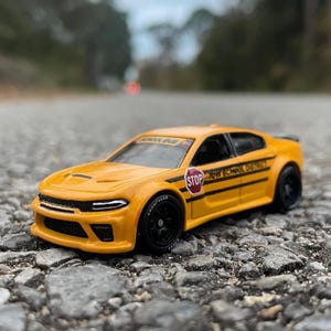May include: A yellow toy car with black wheels and the text "TOW SCHOOL DISTRICT" and a stop sign on the side. The car is on a gray, textured surface. The car has the Goodyear logo on the tire.