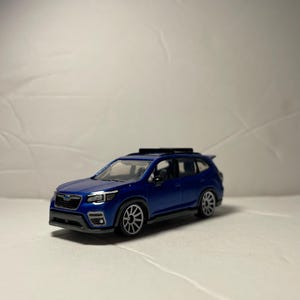 May include: A blue die-cast toy SUV featuring a black roof rack, silver multi-spoke wheels, and black trim along the lower body. The vehicle has clear windows, a detailed front grille with a small emblem, and a compact, sporty design.