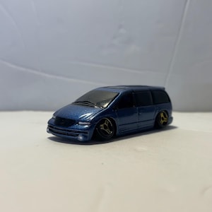 May include: A metallic blue toy minivan with a lowered suspension and custom gold-colored three-spoke wheels. The vehicle features dark tinted windows, a sleek aerodynamic body design, and detailed front headlights and grille elements.
