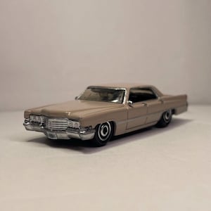 Puede incluir: Un coche de modelo fundido a presión de color beige con detalles cromados. El coche clásico tiene una parrilla delantera detallada, neumáticos negros y un diseño elegante y vintage. El coche de modelo es una réplica en miniatura de un vehículo clásico.