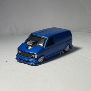 May include: A blue toy van with a custom engine and a black grill. The van has a lowered suspension and white wheels. The vehicle is a miniature model, likely a collectible or a toy car.