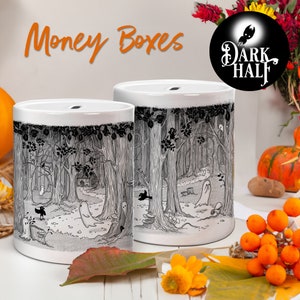 Puede incluir: Dos huchas de cerámica blanca con una escena de bosque en blanco y negro con fantasmas y una casita. El texto "Money Boxes" está en naranja sobre las cajas. El logotipo de The Dark Half está en la esquina superior derecha.