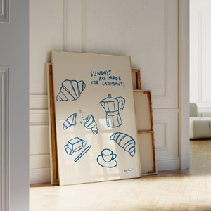 May include: A white framed print with blue illustrations of croissants, butter, a coffee pot, and a cup of coffee. The text on the print reads "Sundays are made for croissants".