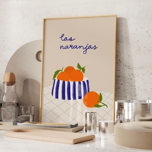 May include: A beige framed print with the text "las naranjas" in blue cursive. The print features a blue and white striped bowl filled with orange oranges and a single orange on the table below the bowl.