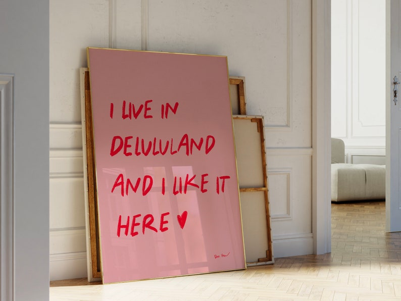 以下が含まれることがあります： 赤いハートのシンボルが付いた赤い文字で「I live in Delulu land and I like it here」と書かれたピンクの額縁入りポスターです。