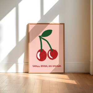 K&ouml;nnte beinhalten: Ein rosa gerahmtes Poster mit einer Zeichnung von zwei roten Kirschen mit gr&uuml;nen Stielen und Bl&auml;ttern. Der Text "SMALL BOOBS, BIG DREAMS" ist unter den Kirschen gedruckt.