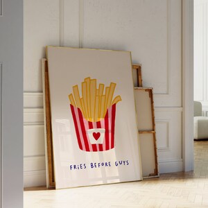 May include: A framed art print featuring a cartoon illustration of a container of french fries. The fries are golden yellow, and the container is red and white striped with a heart. The text "FRIES BEFORE GUYS" is written in blue.