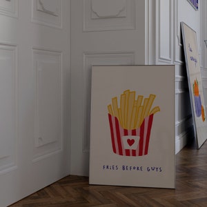 May include: A framed art print featuring a cartoon illustration of a container of french fries with a red and white striped design and a heart. The text "FRIES BEFORE GUYS" is written below the fries. Another art print is leaning against the wall.