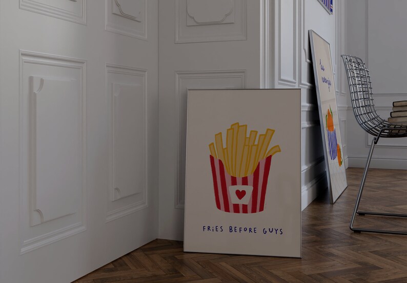 May include: A framed art print featuring a cartoon illustration of a container of french fries with a red and white striped design and a heart. The text "FRIES BEFORE GUYS" is written below the fries. Another art print is leaning against the wall.