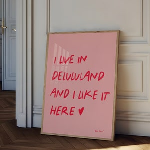 以下が含まれることがあります： 赤い文字で「I live in Delululand and I like it here &hearts;」と書かれたピンク色の額縁入りポスターです。
