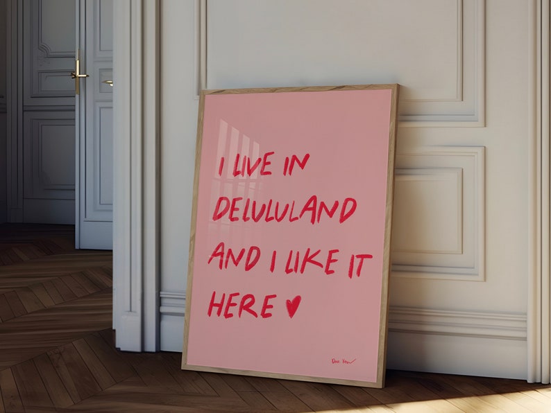 以下が含まれることがあります： 赤い文字で「I live in Delululand and I like it here &hearts;」と書かれたピンク色の額縁入りポスターです。