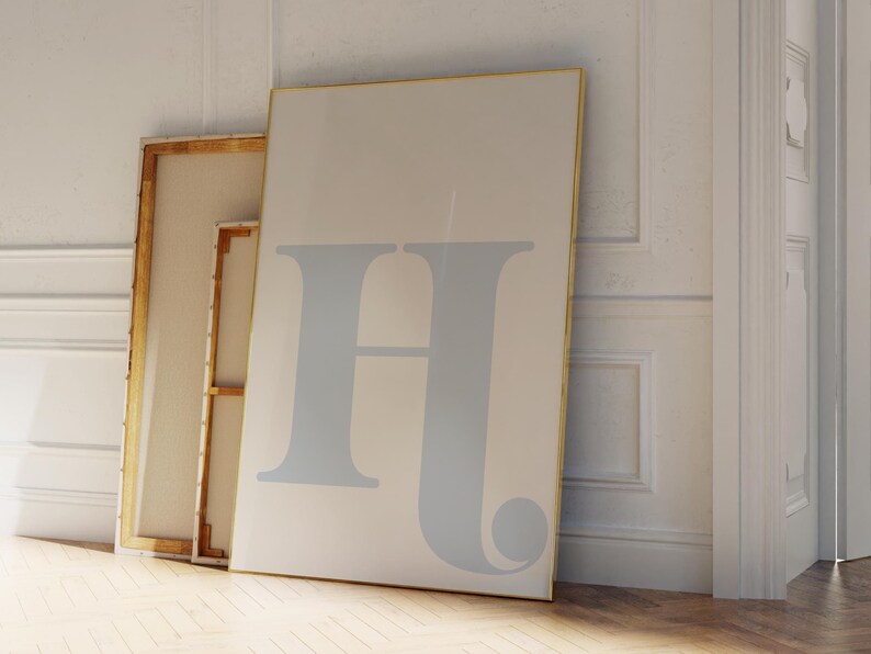 May include: Two framed canvases leaning against a white wall. The larger canvas features a light blue letter "H" in a stylised font. The smaller canvas is wooden and has a white surface.