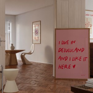 以下が含まれることがあります： 赤い文字で「I live in Delululand and I like it here &hearts;」と書かれたピンク色の額縁入りポスター。ポスターは、木製の床と木製のテーブルと椅子のある部屋の壁にかけられています。