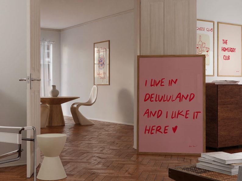 以下が含まれることがあります： 赤い文字で「I live in Delululand and I like it here &hearts;」と書かれたピンク色の額縁入りポスター。ポスターは、木製の床と木製のテーブルと椅子のある部屋の壁にかけられています。