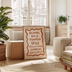 May include: A framed print with a white background and brown text that reads "Gilmore Girls Pumpkin Spice Mulled Wine Candles Cozy Blankets". The print is leaning against a cardboard box in a room with a white couch and a window.