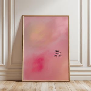 Puede incluir: Una impresión artística enmarcada con un diseño abstracto en tonos rosa, melocotón y amarillo. El texto escrito a mano dice "HOW LUCKY ARE WE?". El marco es de color madera clara. La obra de arte se exhibe contra una pared blanca.