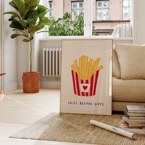 May include: A framed art print featuring a cartoon illustration of golden french fries in a red and white striped container with a heart. The text "FRIES BEFORE GUYS" is below the fries. The print is leaning against a wall in a living room.