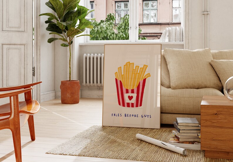 May include: A framed art print featuring a cartoon illustration of golden french fries in a red and white striped container with a heart. The text "FRIES BEFORE GUYS" is below the fries. The print is leaning against a wall in a living room.