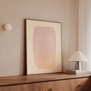 May include: A framed abstract art print featuring a large, vertical shape with a gradient of pink and peach hues. The artwork is displayed on a wooden cabinet, with a white lamp and books nearby. The overall aesthetic is minimalist and modern.