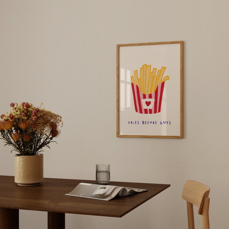 May include: A framed art print featuring a cartoon illustration of a container of french fries. The fries are yellow, and the container is red and white striped with a heart. The text "FRIES BEFORE GUYS" is below the image.