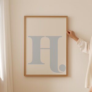 May include: A framed print featuring a large, light blue letter "H" in a simple, modern font. The frame is a light wood colour.