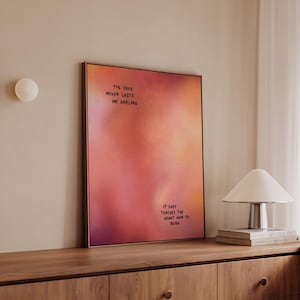 Könnte beinhalten: Ein rechteckiges gerahmtes Wandbild mit einem Farbverlauf in gemischten Rosa-, Orange- und Violetttönen. Der Text lautet: THE COLD NEVER LASTS MY DARLING und IT JUST TEACHES THE HEART HOW TO BURN in einer einfachen, dunklen, handschriftlichen Schrift.