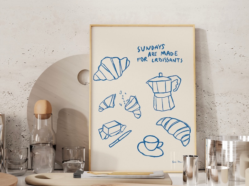 Op de afbeelding: Een blauwe en witte illustratie van een koffiepot, croissants, boter en een kopje koffie. De tekst luidt "Zondagen zijn gemaakt voor croissants".