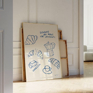 Op de afbeelding: Een wit ingelijst print met blauwe illustraties van croissants, boter, een koffiepot en een kopje koffie. De tekst op de print luidt "Zondagen zijn gemaakt voor croissants".