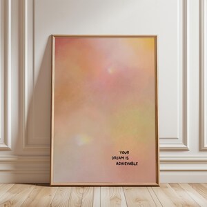 Könnte beinhalten: Ein gerahmter Kunstdruck mit einem Hintergrund im Aquarellstil in Rosa-, Pfirsich- und Gelbtönen. Der schwarze Text "YOUR DREAM IS ACHIEVABLE" ist unten rechts aufgedruckt. Der Rahmen ist hellbraun.