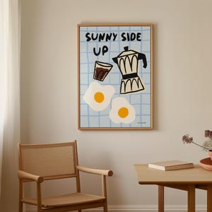 May include: A framed illustration of a sunny side up egg, a cup of coffee, and a coffee pot with the text "Sunny Side Up" in black lettering on a blue and white checkered background.