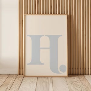 May include: A framed print featuring a light blue letter H on a cream background. The frame is a light wood colour.