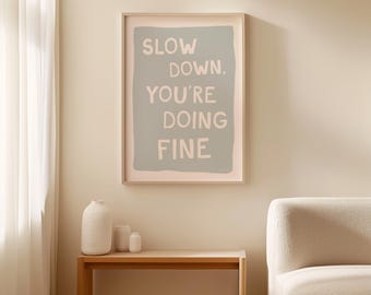 May include: A framed print with a light blue background and white text that reads "Slow down, you're doing fine". The print is hanging on a light beige wall above a wooden table with a vase and two small jars.