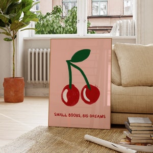 K&ouml;nnte beinhalten: Rosa-wei&szlig;er gerahmter Kunstdruck mit einer Zeichnung von zwei roten Kirschen mit gr&uuml;nen Stielen und Bl&auml;ttern. Der Text "SMALL BOOBS, BIG DREAMS" ist unter den Kirschen gedruckt.
