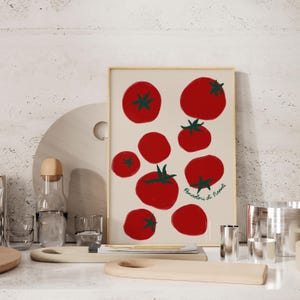 May include: A framed art print featuring a beige background with red tomato illustrations and green stems. The print includes the text "Pomodori di Napoli" in green script. The artwork is displayed on a white surface with kitchen accessories.
