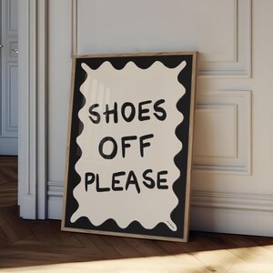 Può includere: Una stampa incorniciata in bianco e nero con uno sfondo bianco e testo nero che recita "SHOES OFF PLEASE" in un font ondulato disegnato a mano.