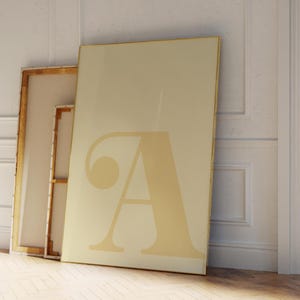 May include: Three framed art pieces leaning against a white wall. The largest canvas features a large, stylized letter "A" in a light beige colour. The other two canvases have a similar colour scheme and are leaning against the wall.