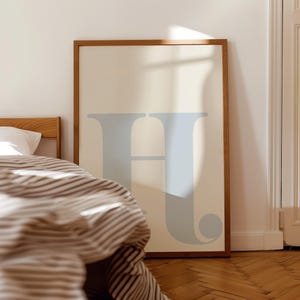 May include: A framed print featuring a light blue letter "H" on a cream background. The print is hanging on a white wall above a bed with a striped duvet.