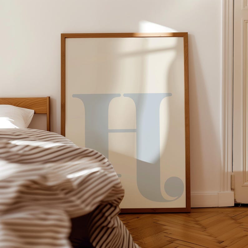 May include: A framed print featuring a light blue letter "H" on a cream background. The print is hanging on a white wall above a bed with a striped duvet.